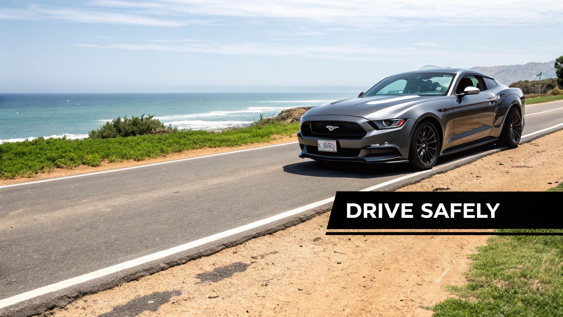 A sleek grey Ford Mustang parked on a scenic coastal road overlooking the ocean under a clear sky.